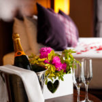 Close up shot of champagne and flowers left in a privilege room when dressed as a bridal suite. Out of focus behind the champagne is the four poster bed topped with red rose petals.