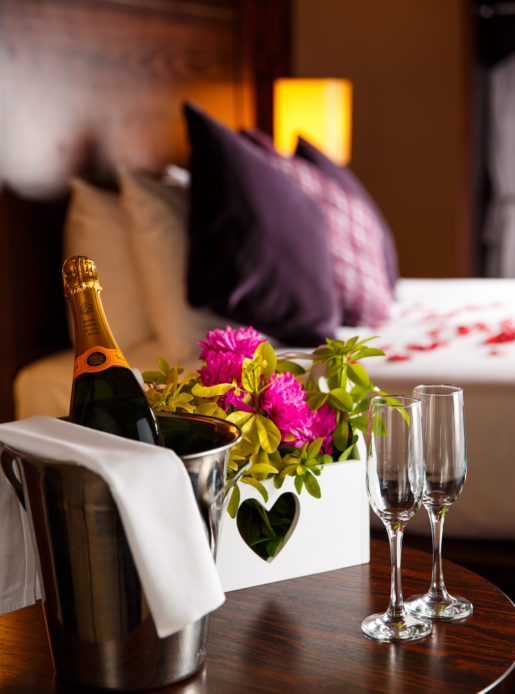 Close up shot of champagne and flowers left in a privilege room when dressed as a bridal suite. Out of focus behind the champagne is the four poster bed topped with red rose petals.